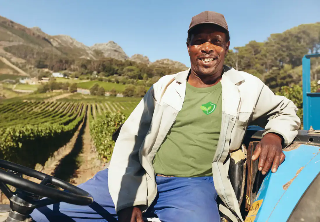 A smiling farm worker in a Crop Compliance shirt on a tractor beside a vineyard